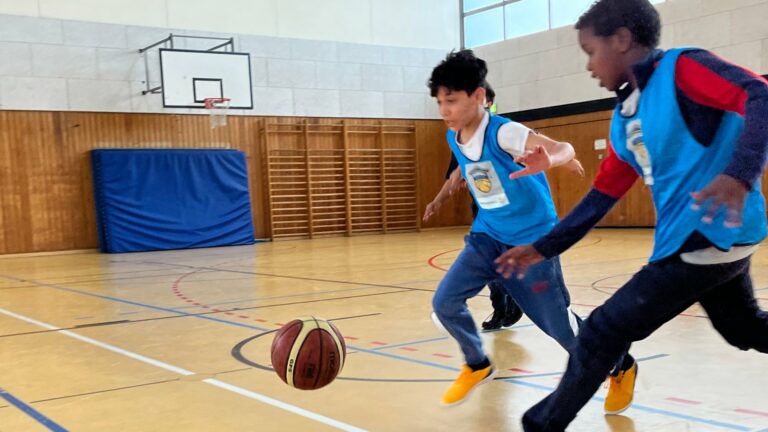 Zwei Jungen rennen in einer Turnhalle mit Trikot einem Basketball hinterher.