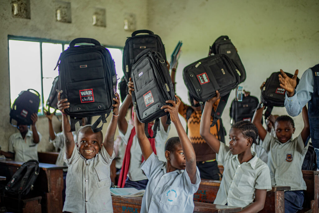 In einem vollen Klassenzimmer halten Kinder stolz ihre Rucksäcke hoch