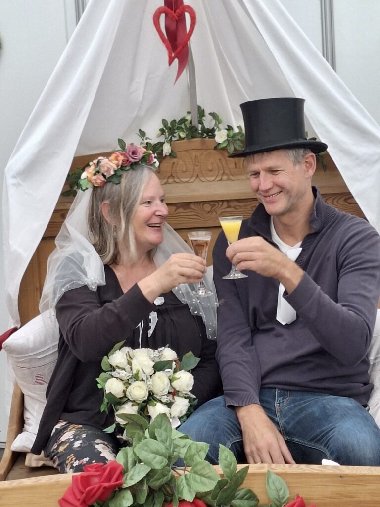 Ein Ehepaar sitzt gemeinsam vor einem Klavier und stoßt in feierlicher Kleidung an.