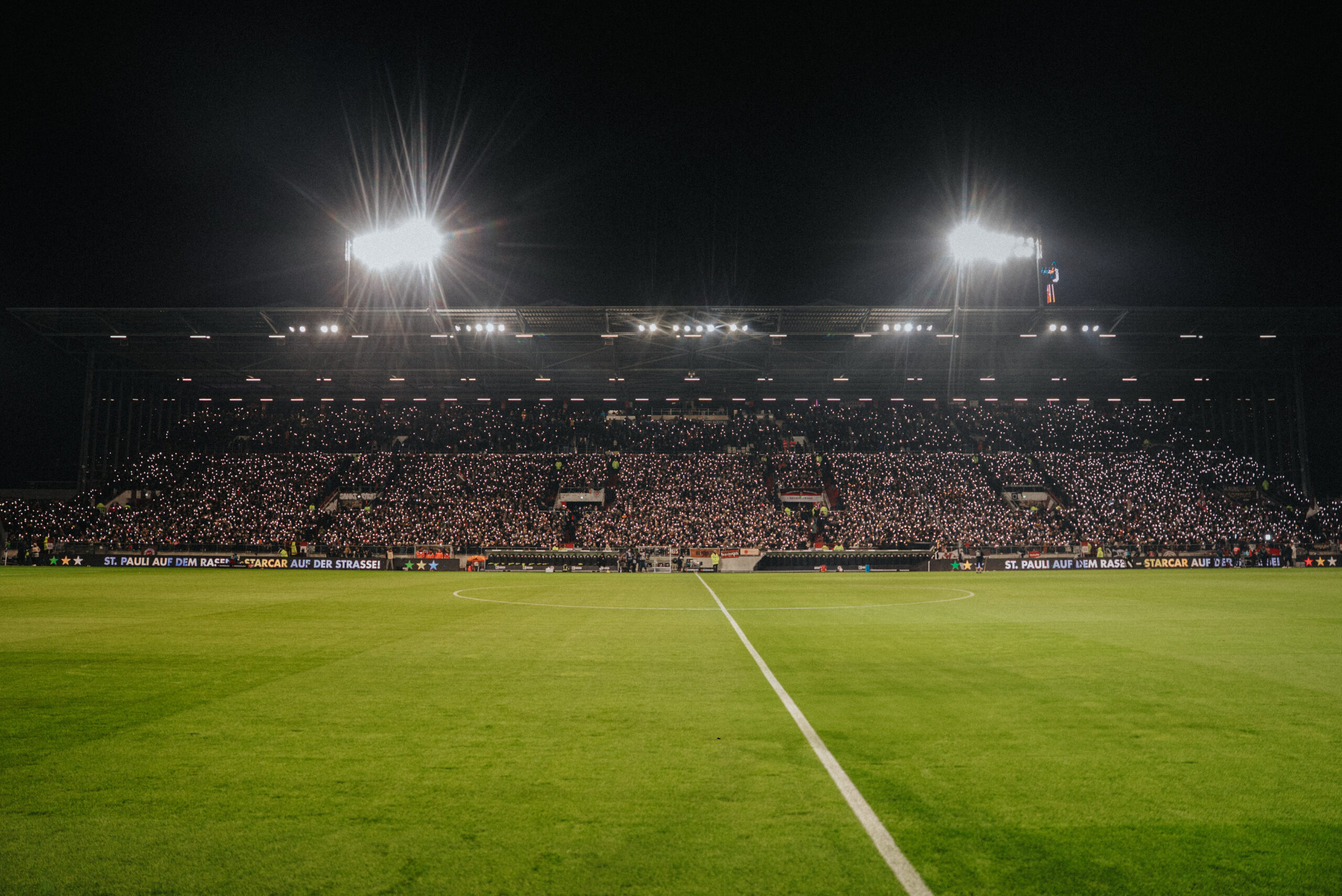 Rasen eines Fußballstadions, dahinter die Tribüne, die von vielen kleinen Lichtern übersät ist