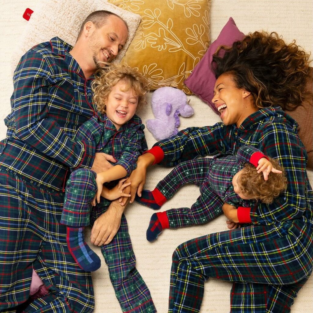 Ein Paar liegt gemeinsam mit zwei Kleinkindern auf einem großen Bett. Alle tragen blaukarierte Pyjama.