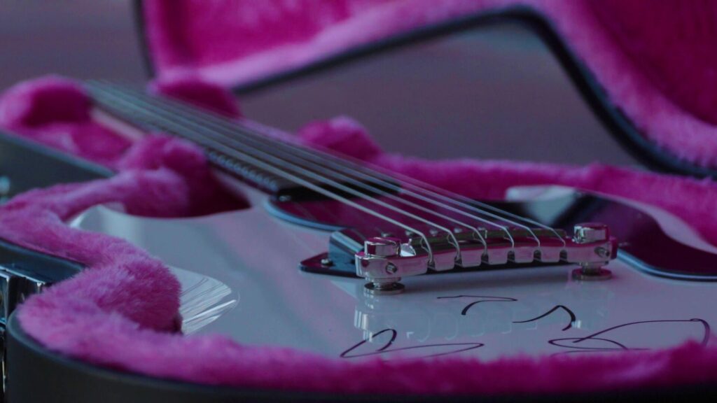 Weiße Gitarre mit schwarzer Unterschrift in einem pinken, offenen Gitarrenkoffer.