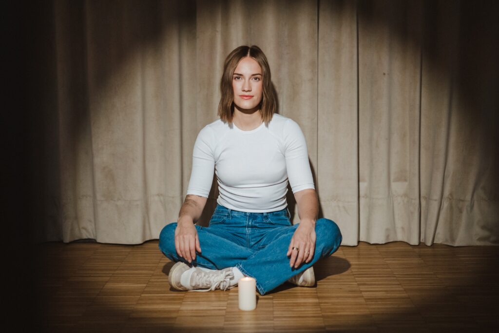 Frau sitzt im Schneidersitz auf dem Boden und blickt in die Kamera. Vor ihr steht eine Kerze.