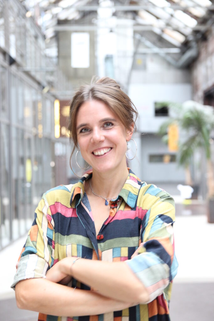 Eine brünette Frau mit hochgesteckten Haaren und einem bunten Hemd, lächelt mit verschränkten Armen in die Kamera.