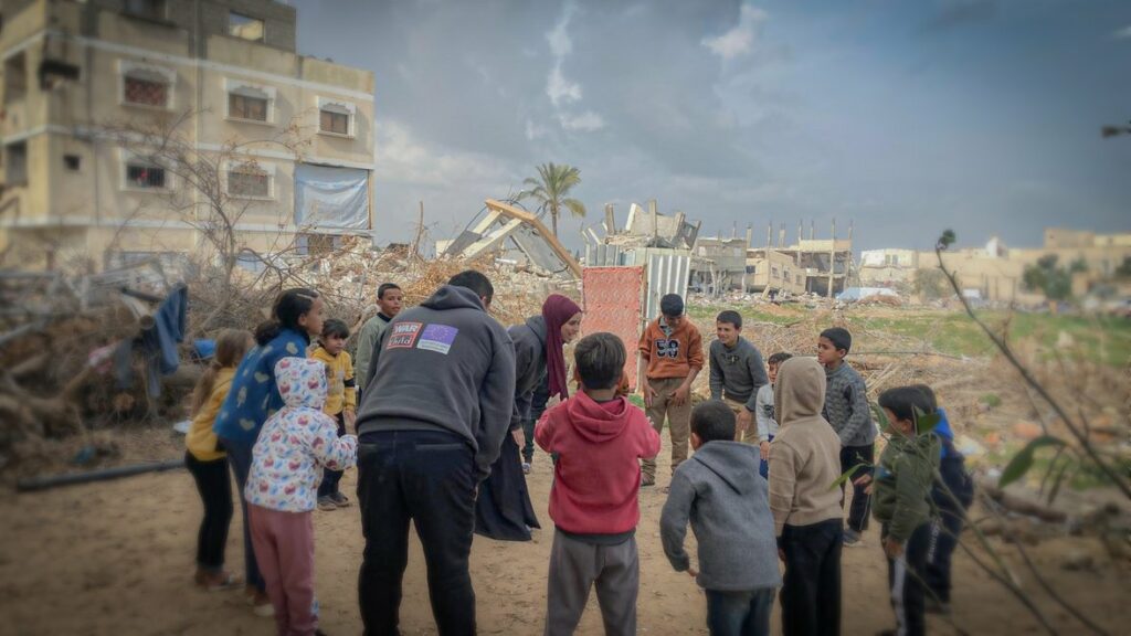 Eine Gruppe von Kindern steht gemeinsam mit zwei Erwachsenen, die einen War-Child-Pullover tragen, im Kreis. Um sie herum befinden sich Trümmer von Gebäuden.