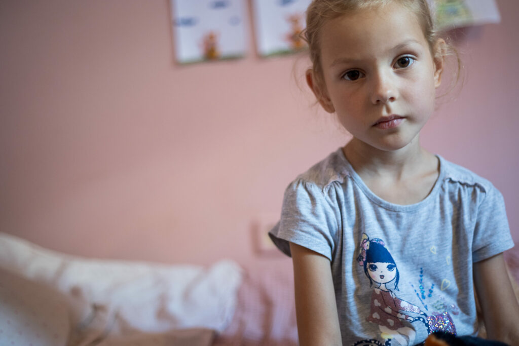 Blondes Mädchen im Alter von etwa 5 Jahren schaut trostlos. Sie sitzt in einem Rosa gestrichenen Zimmer auf ihrem Bett.