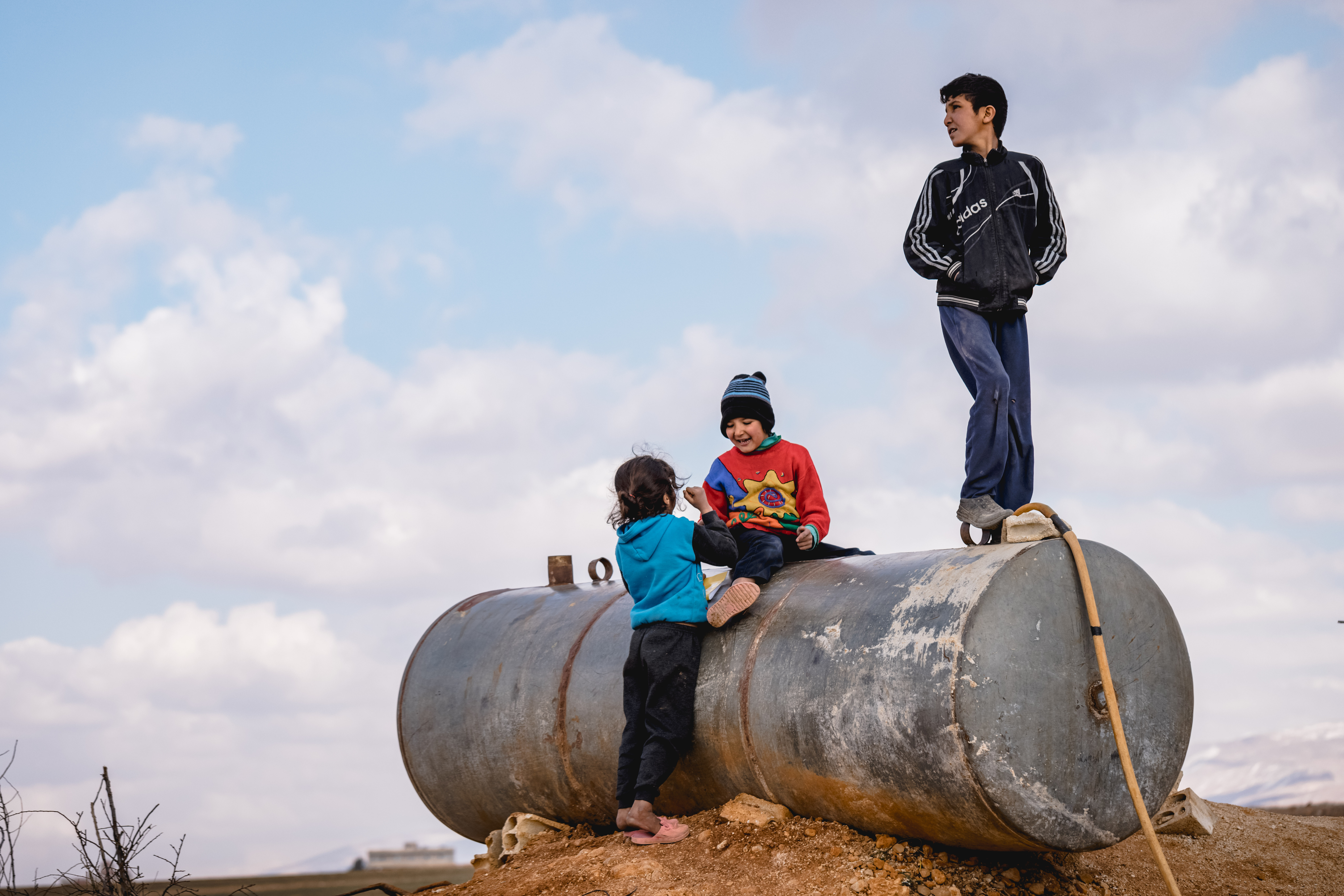 Drei Kinder spielen auf und an einem großen, verwitterten Metalltank, der auf einem Erdhügel in einer weiten, ländlichen Landschaft unter leicht bewölktem Himmel steht.