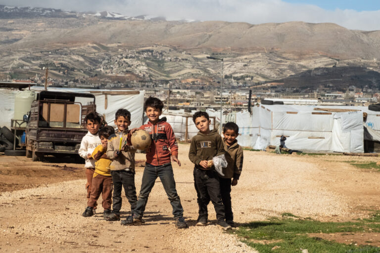 eine Gruppe von fünf kleinen Jungs steht lachend vor einem Flüchtlingslager. EIner von ihnen hat einen Fußball in der Hand.
