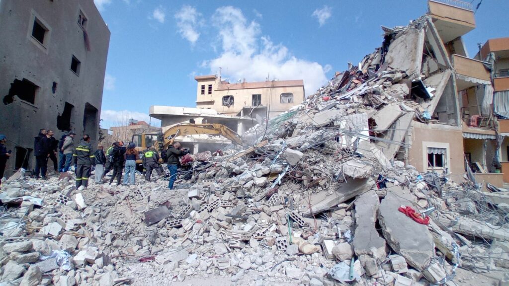 Neben einem zerbombten Hausblock ist eine Gruppe von Einsatzkräften die auf den Trümmern steht.
