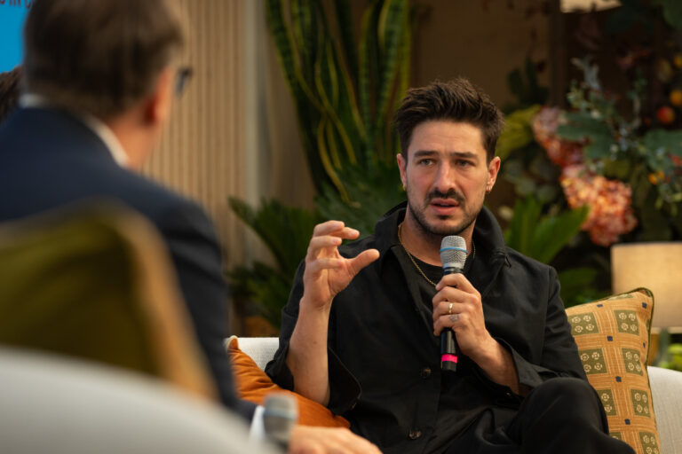 Marcus Mumford hält ein Mikrofon und sitzt auf einem Sofa. Er spricht mit jemandem, der mit dem Ruücken zur Kamera gekehrt ist .